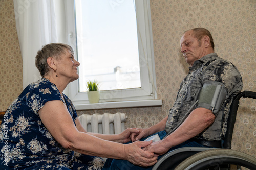 an elderly woman measures the pressure of her elderly spouse who sits in a wheelchair while holding his hands and gently looks into his eyes