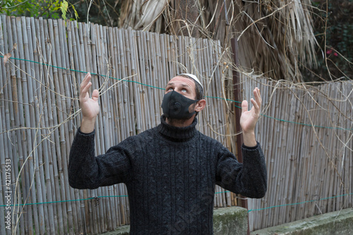 Jewish man wearing kippah in Hebrew or yarmulke (cloth cap traditionally worn by Jewish males) with mask against Corona virus Looking And Holding His Hands Up To The Sky praying and looking up for God photo