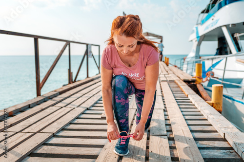 An adult woman in sportswear, sitting down tying her shoelaces. In the background, the pier with the ship and the sea. The concept of sport and a healthy lifestyle © _KUBE_