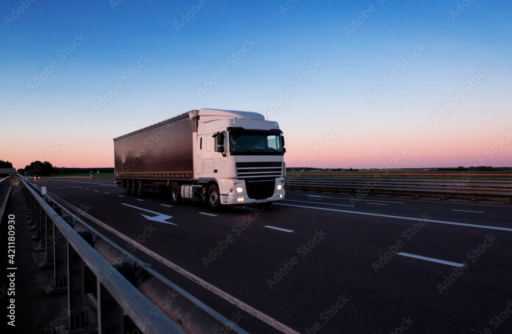 A white semi-trailer truck drives down the highway in the evening with its headlights on. Blue sky and sunset. The mode of work and rest of truck drivers. Copy space for text