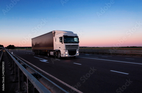 A white semi-trailer truck drives down the highway in the evening with its headlights on. Blue sky and sunset. The mode of work and rest of truck drivers. Copy space for text