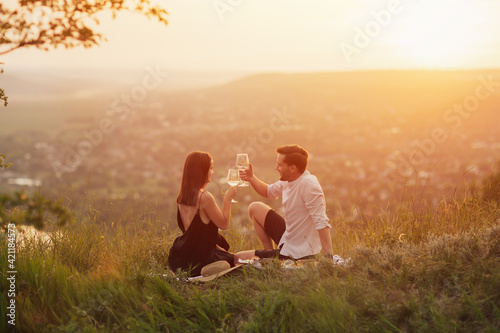 Attractive couple sitting on a picnic on the hill and looking at each other while clinking with glasses of wine. Happy young couple is relaxing at nature. 