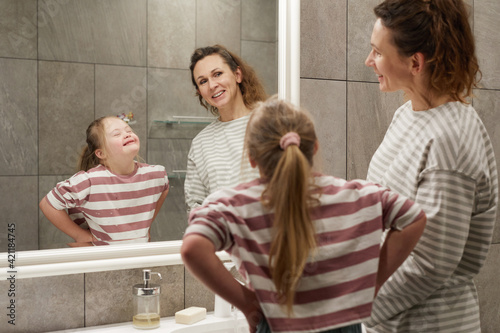 Portrait of cute little girl with down syndrome making faces at mirror while having fun with mother in bathroom, copy space