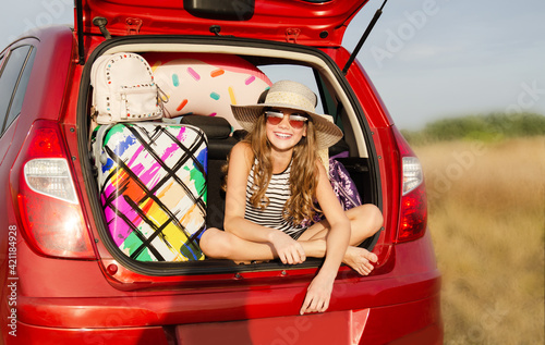 Family ready for the travel on summer vacation. Cute happy smiling little girl child is sitting in the red car with luggage and bags outdoors.