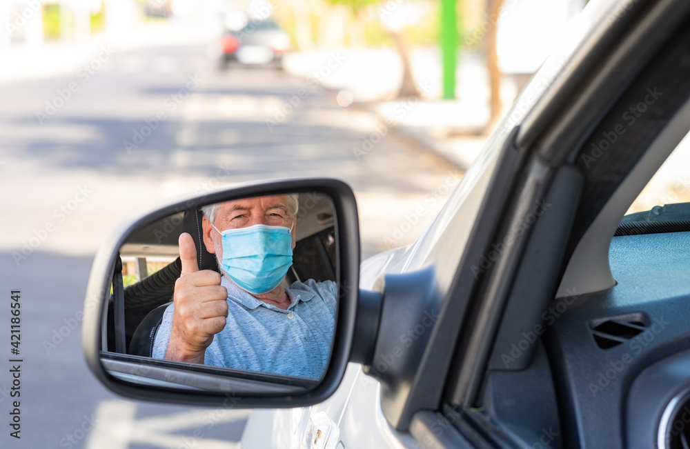 Elderly man wearing a protective mask while driving the car looking at the camera in the mirror
