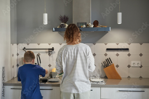 Back view portrait of little boy washing dishes in kitchen while helping mother  copy space