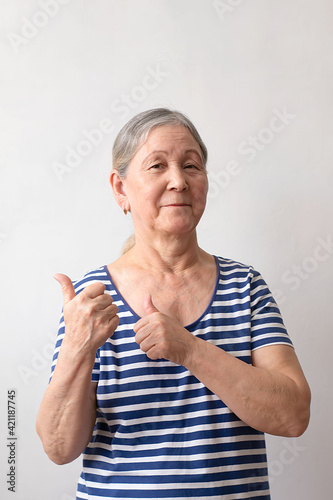 Elderly older woman, gray-haired lady of the 60s in a striped white and blue dress pointing with her hand to her back and thumbs up on a white background