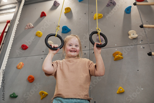 Fototapeta Naklejka Na Ścianę i Meble -  Waist up portrait of cute girl with down syndrome enjoying exercise at home or at sports center and looking at camera while holding at rings against climbing wall, copy space
