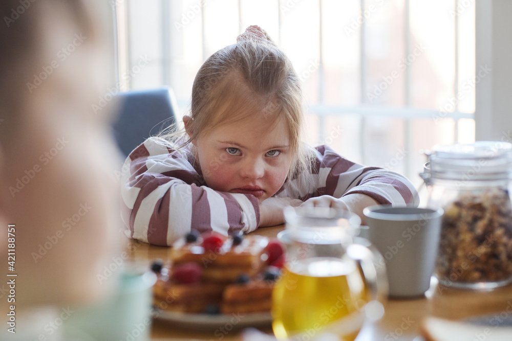 Portrait of frustrated teenage girl with down syndrome looking at camera while sitting at table during breakfast in kitchen, copy space