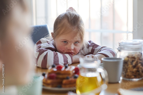 Portrait of frustrated teenage girl with down syndrome looking at camera while sitting at table during breakfast in kitchen, copy space photo