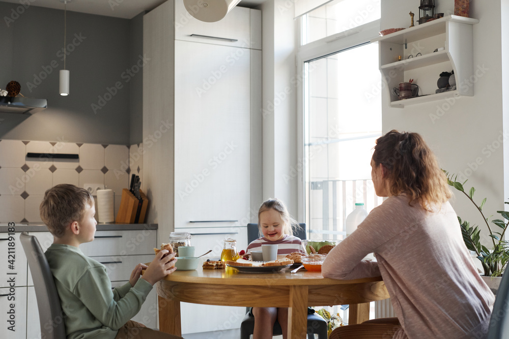 Wide angle view at happy girl with down syndrome enjoying breakfast with family at home while sitting at wooden table together, copy space Wide angle view at happy girl with down syndrome enjoying breakfast with family at home while sitting at wooden table together, copy space