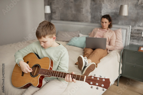 Portrait of little boy playing guitar while sitting on bed with mother using laptop in background, copy space