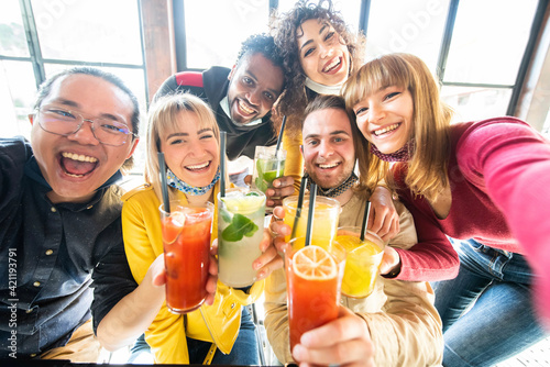 Multiracial people wearing protective face masks drinking cocktails at bar restaurant - New normal friendship concept with young friends taking a selfie - Food, drink and people concept.