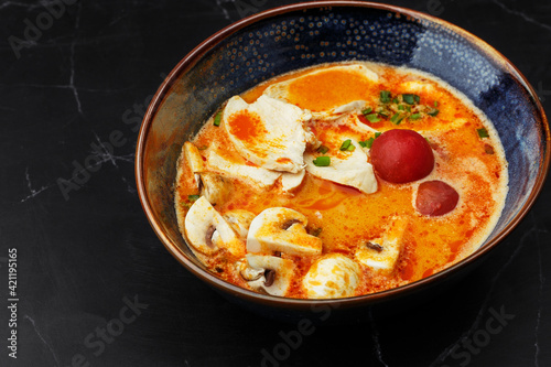 Asian chicken curry soup with chicken breast, tomatoes, mushrooms and spring onion. Close-up photo of the dish served in a dark blue bowl, isolated on a black marble background. Asian cuisine.