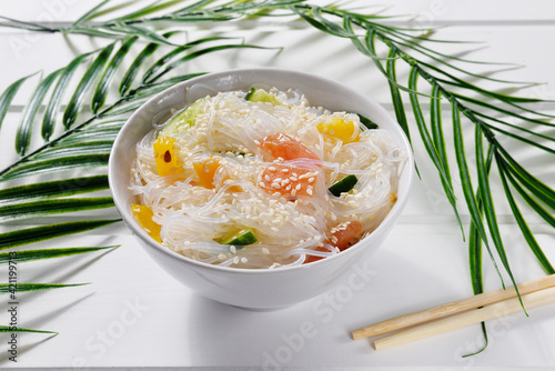 Chinese glass funchosa noodles with vegetables and sesame seeds on a white wooden background with palm leaves. photo