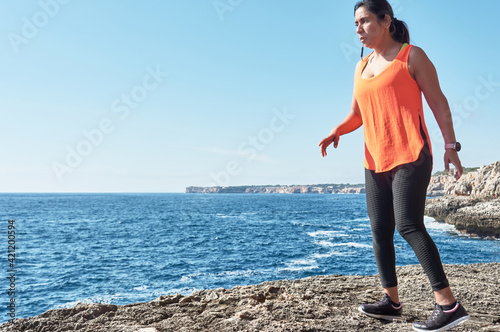 Fototapeta Naklejka Na Ścianę i Meble -  Latin woman, middle-aged, wearing sportswear, training, doing physical exercises, plank, sit-ups, climber's step, burning calories, keeping fit, outdoors by the sea, wearing headphones, smart watch