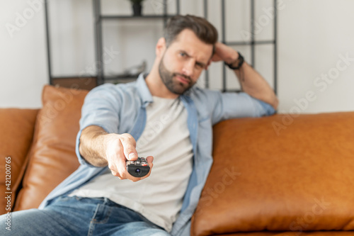 Blurred image of a mad young bearded hipster guy sitting on the couch in the living room, pointing a tv remote on the camera, switching channels, unhappy with wifi issues or television program