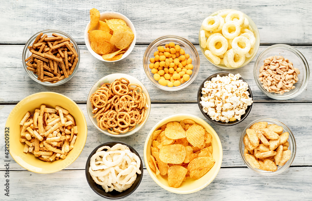 Assorted chips, popcorn, onion rings, croutons, cheese balls, cookies in bowls. unhealthy nutrition for figure, heart, skin, teeth. An assortment of fast carbohydrates. Junk food on wooden
 background