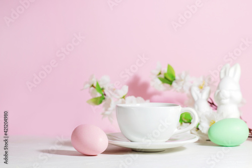 White mug with tea or coffee near pink and green Easter eggs, rabbits and flowers, on a concrete white table, on a pink background, with hard shadows. Easter breakfast concept.