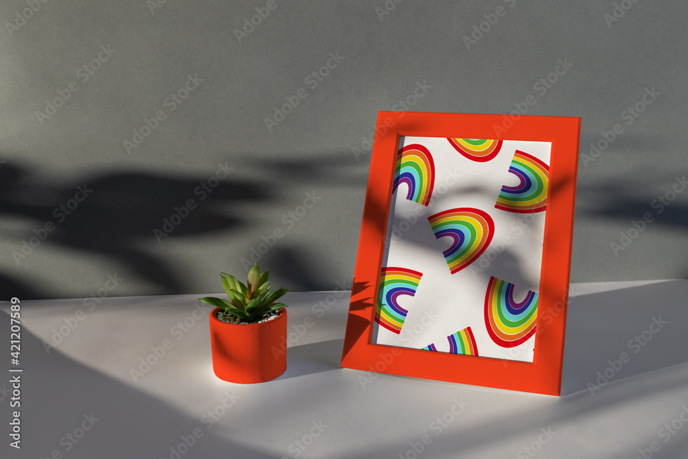 Obraz premium Dark flat lay. Red frame with rainbow photo and small flower pot near on a textured white paper background. Natural light casts a shadow from the plants.