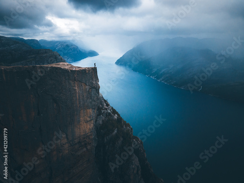 A man with a blue jacket, shorts, hiking boots, and backpack standing on the top of the cliff Preikestolen and overlooking a fjord in Norway. Moody landscape and sky. © finnghal