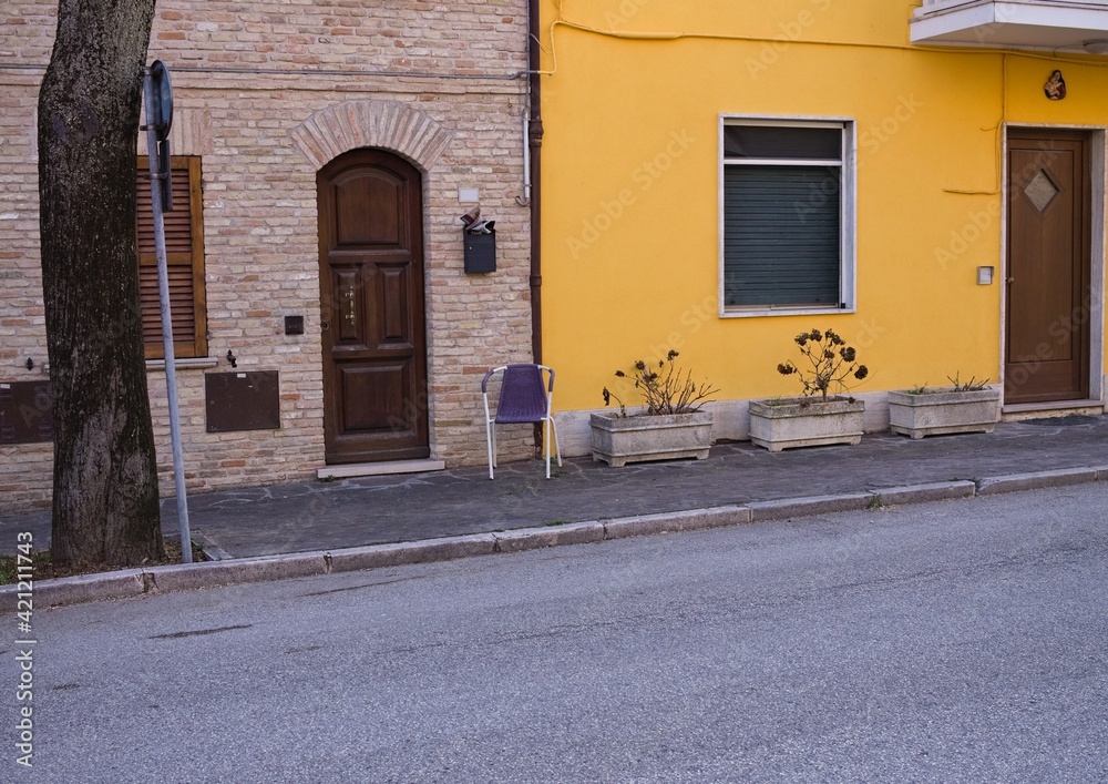 An isolated chair on the sidewalk in front of a yellow and brick house (Marche, Italy, Europe)
