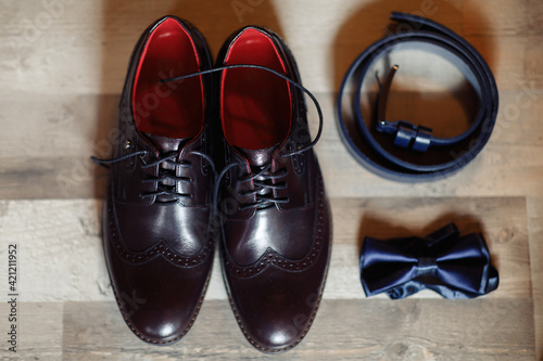 Leather classic brown shoes with a red insole on a wooden floor near the belt and bow tie. Set of gentleman and groom for the wedding.