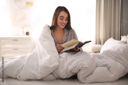Beautiful young woman wrapped with soft blanket reading book on bed at home