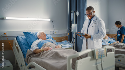 Hospital Ward  Friendly Black Doctor Talks with Elderly Caucasian Patient Resting in Bed. Doctor Uses Tablet Computer  Does Checkup. Old Man Fully Recovering after Successful Surgery