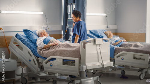 Hospital Ward: Friendly Female Head Nurse Making Rounds does Checkup on Elderly Patient Resting in Bed. She Monitors Vitals while Old Man Fully Recovering after Successful Surgery