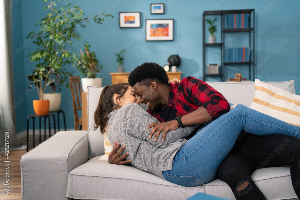 Close up of the cheerful young Multi-ethnic couple lying on the couch, hugging and laughing. Inside. Close up of the cheerful young Multi-ethnic couple lying on the couch, hugging and laughing. Inside.