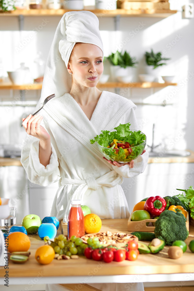 Adorable woman in bathrobe and towel eating salad, standing with lots of healthy fresh food in kitchen Adorable woman in bathrobe and towel eating salad, standing with lots of healthy fresh food in kitchen