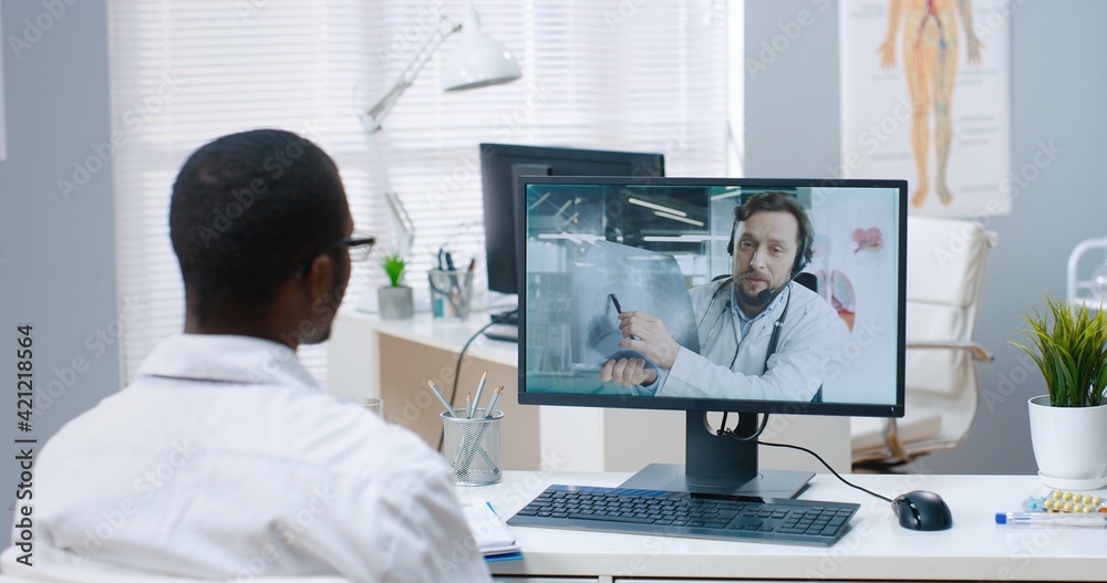 Over shoulder view of African American male physician speaking on online video call on computer with Caucasian man doctor colleague sitting in hospital, specialist consulting on disease treatment
