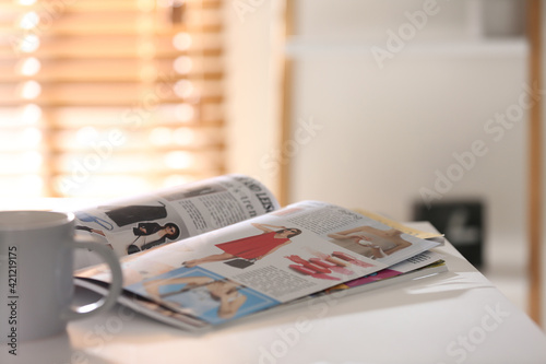 Fashion magazines and cup of hot drink on white table indoors