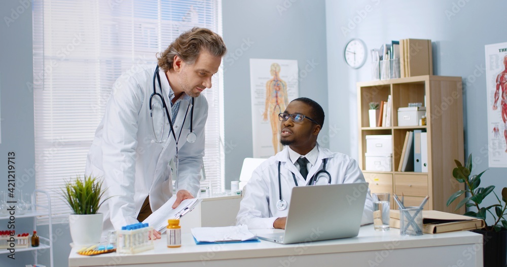 Caucasian man doctor stands near African American young physician in cabinet in clinic speaking and consulting tapping on computer looking at screen and discussing disease treatment healthcare workers
