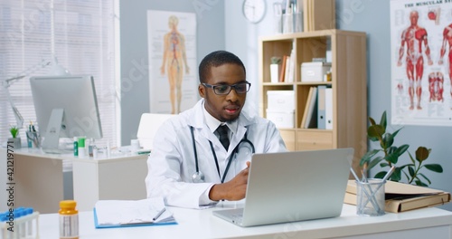 Portrait of young handsome African American male doctor in white coat videochatting on laptop sitting in clinic in cabinet having online video consultation with patient. Healthcare, disease treatment
