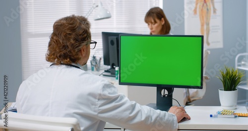 Over shoulder view of Caucasian male physician sitting in hospital cabinet and looking at green screen on computer writing in notebook. Medical consultation, video call with doctor, chroma key photo