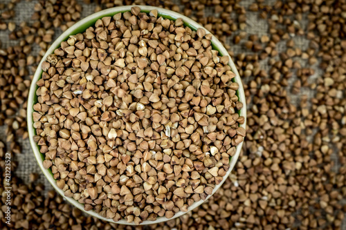 Buckwheat is poured in a round bowl (plate), close-up top view, around defocused buckwheat scattered around the table