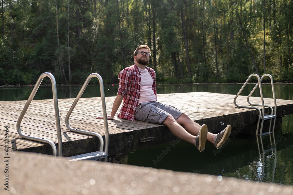 Calm man sits on the edge of a wooden pier and looks up thoughtfully, against the background of a calm forest lake. Lifestyle. Calm man sits on the edge of a wooden pier and looks up thoughtfully, against the background of a calm forest lake. Lifestyle.