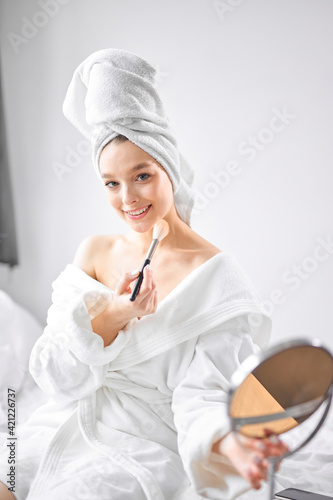 Good-looking happy woman with towel on head, holding mirror and applying make up with brush
