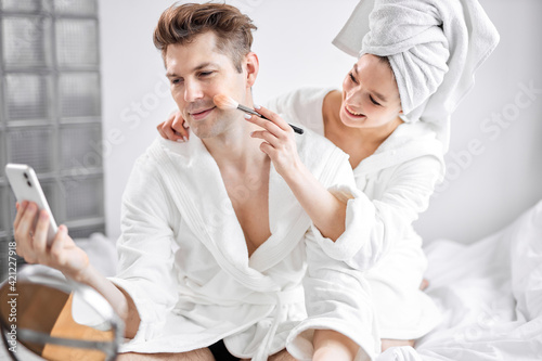 cheerful young caucasian couple in bathrobes and towel have fun, woman doing make-up on boyfriend's face