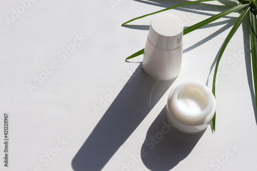 Face cream in glass jar, bottle of lotion and palm leaves on white table background. Hard light, shadow. Natural beauty products for skin and body care. Summer lifestyle. Top view, copy space	