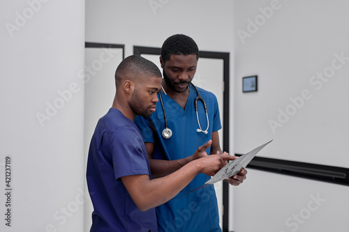 Two african american male colleagues doctors working together, discussing and looking at clipboard on clinic.
