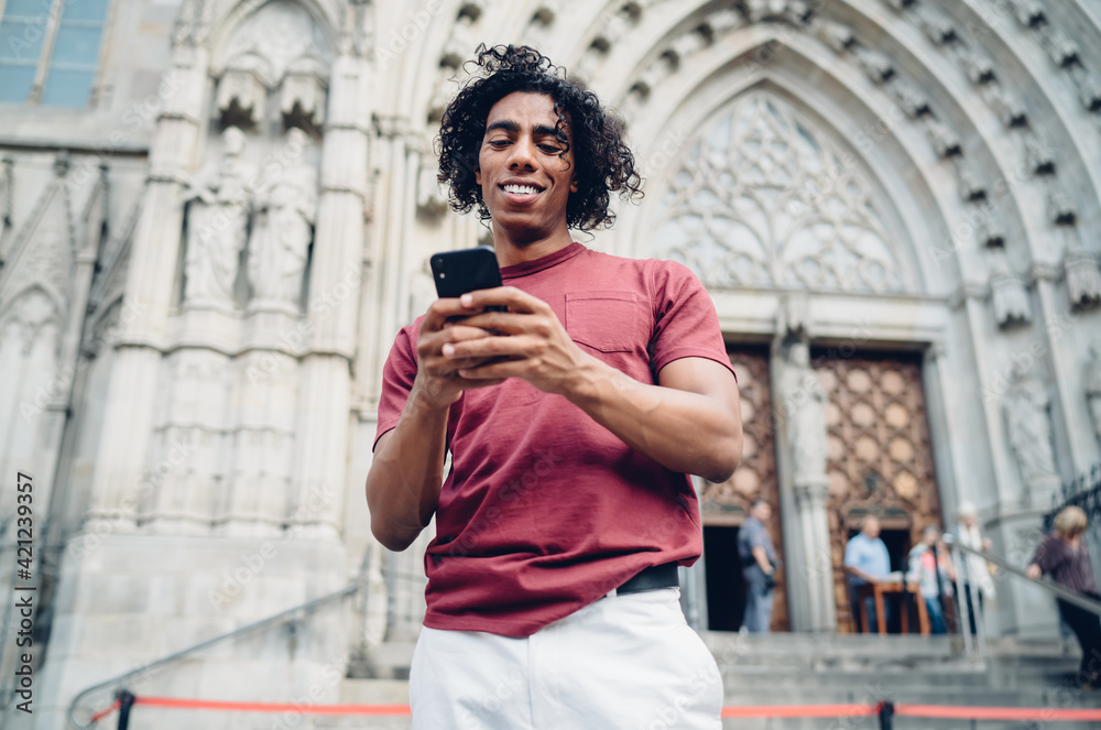 Happy travel blogger with curly hair using cellphone gadget for social networking during journey vacations for visiting historic city, smiling hipster guy connecting to 4g internet for browsing