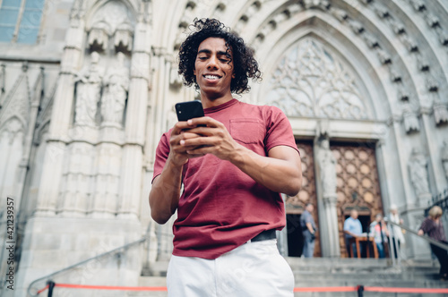 Happy travel blogger with curly hair using cellphone gadget for social networking during journey vacations for visiting historic city, smiling hipster guy connecting to 4g internet for browsing