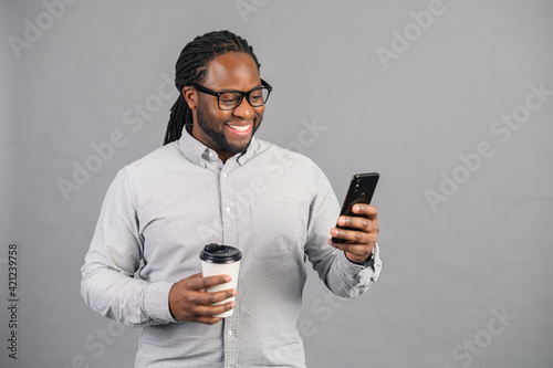 Busy and confident African-American businessman wearing smart casual shirt and eyeglasses texting on the smartphone and holding a paper cup of take-away coffee, male office employee isolated on grey