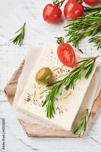 Cheese feta with rosemary, herbs, cherry tomatoes, olives and olive oil on wooden cutting board on white old wooden background. Traditional Greek homemade cheese. Selective focus.