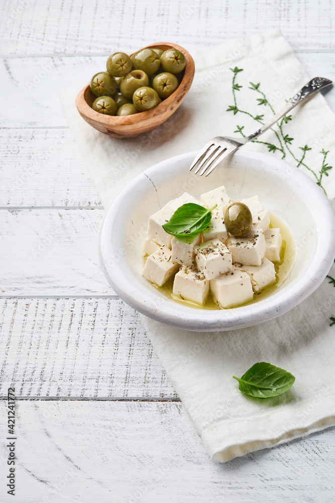 Feta cheese cubes with rosemary, olives and olive oil sauce in white bowl on light gray background. Traditional Greek homemade cheese. Selective focus.