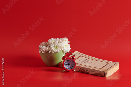 white flowers in a green cup stand next to an alarm clock and a book 
