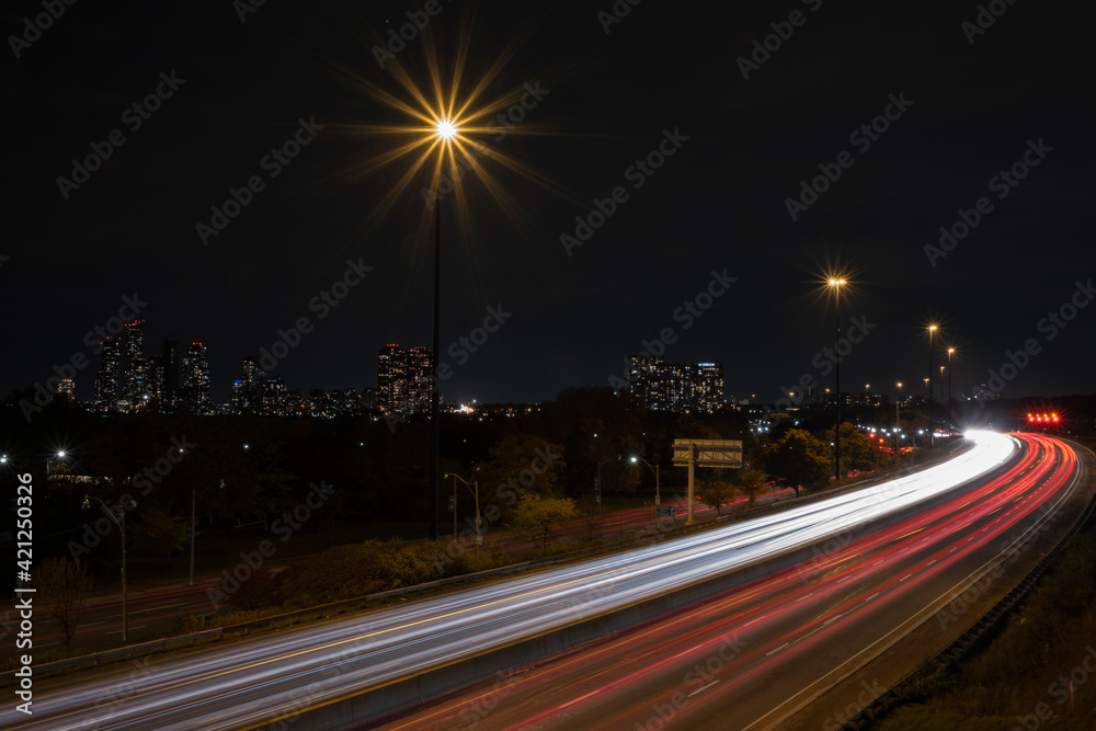Aerial view of highway with fast moving cars creating long light trails, city skyline in the background. High travel speed effect, dui, dangerous, careless, stunt, drunk driving concept. Night scene.